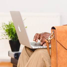 A laptop rests on the knees of a person wearing khaki pants. They are typing on the laptop keyboard. A satchel sits in front of the person's body and a side table with a plant are visible in the background.